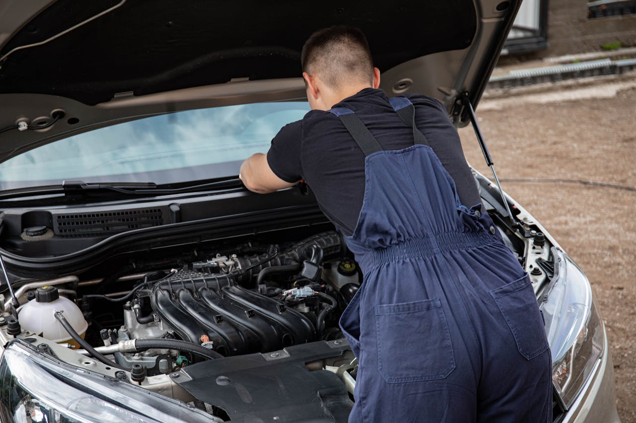 A mechanic in overalls fixing a car engine outdoors on a sunny day.