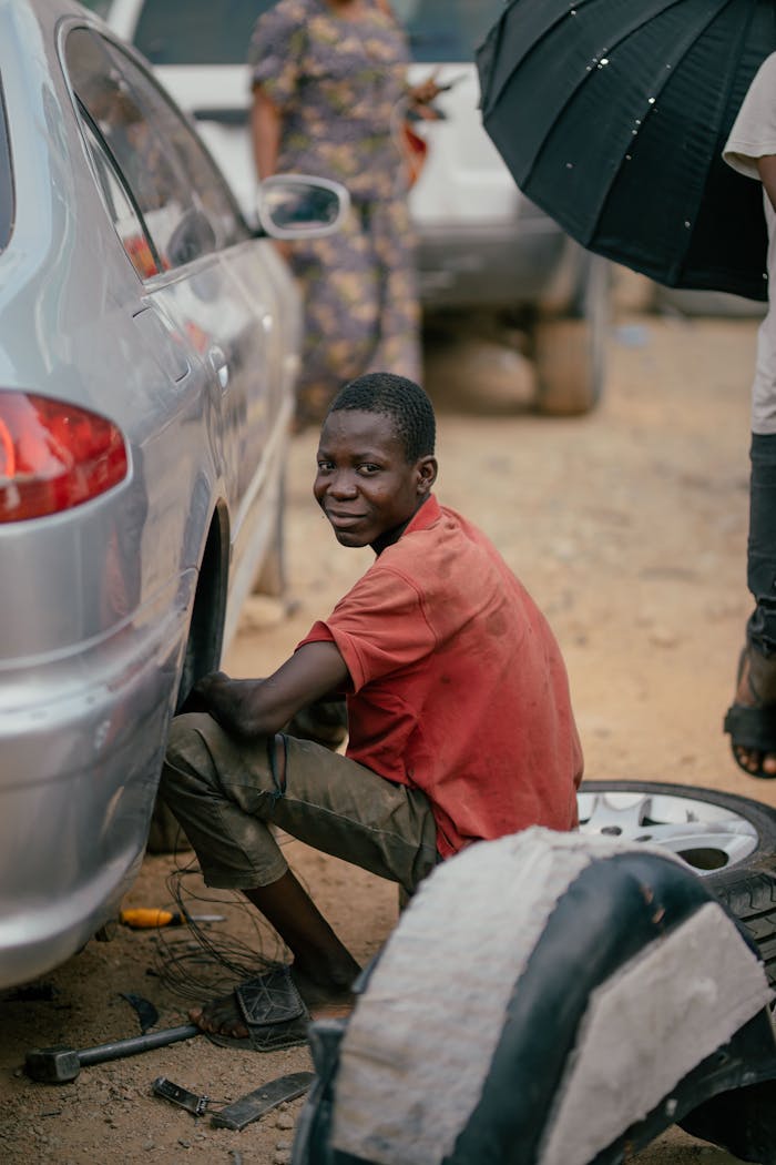 Young mechanic smiling while fixing car tire outside, highlighting craftsmanship and community.