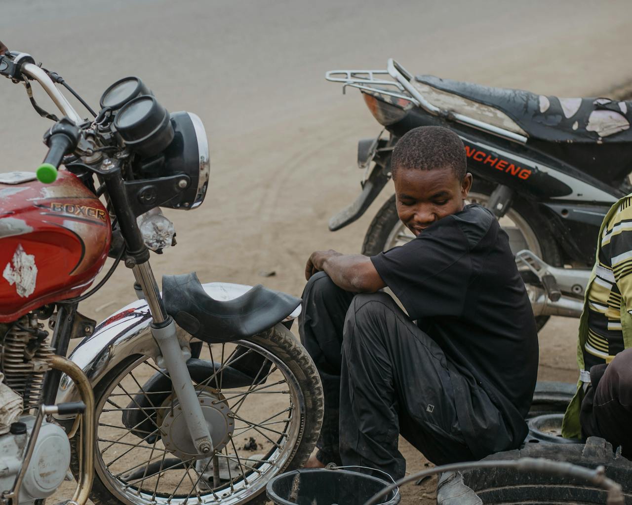 African man sitting beside parked motorcycles, working on repairs in an outdoor setting.